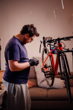 Eldivenli odaklanmış bir adam bisiklet pedalını inceliyor, onu kapalı bir çalışma alanındaki kırmızı bir bisiklete takmaya hazır..
