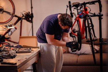 Eldivenli odaklanmış bir adam bisiklet pedalını inceliyor, onu kapalı bir çalışma alanındaki kırmızı bir bisiklete takmaya hazır..