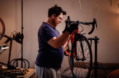 Eldivenli odaklanmış adam rahat bir ev atölyesindeki kırmızı yol bisikletinin gidonunu ve ön tekerlek hizasını kontrol ediyor..