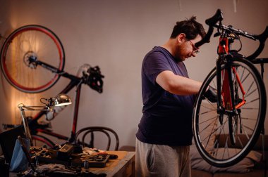 Odaklanmış adam tamir tezgahına monte edilmiş kırmızı bir bisiklet üzerinde çalışıyor. Etrafı, rahat bir çalışma alanındaki aletler ve parçalarla çevrili..
