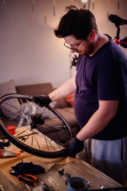 Eldiven giyen adam, rahat bir ev ortamında dağınık bir masa üzerindeki aletler ve parçalarla çevrili bisiklet tekerleğini yakından inceliyor..
