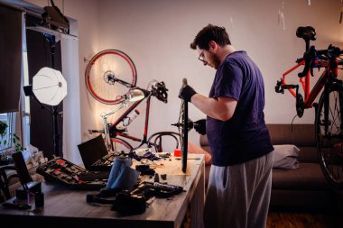 Eldivenli bir adam ev atölyesinde bisiklet tekerleğini inceliyor. Etrafı aletlerle, ışıklarla ve yaratıcı iç mekanlarda birden fazla bisikletle çevrili..