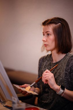Odaklanmış genç bir kadın fırçayla bir palete renk katıyor, kapalı bir çalışma alanında sanat yaratmaya hazırlanıyor..