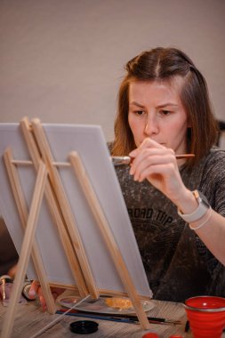 Konsantre olmuş genç bir kadın, bir fırça kullanarak tuvale resim yapıyor, kapalı bir yaratıcı seans sırasında sanatsal çalışmalarıyla yakından ilgileniyor..