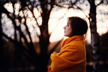 Kadın sıcak turuncu bir battaniyeye sarılmış, gün batımına dikkatlice bakıyor..