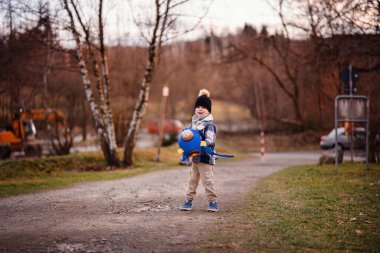 Rahat kışlık giysiler içindeki küçük çocuk dışarıda oyuncak ayıyla mavi bir roket çantası sallıyor..