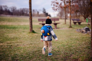Rahat kış kıyafetleri içinde küçük bir çocuk çimenli bir parkta oyuncak ayıyla birlikte mavi bir roket çantasıyla yürüyor..