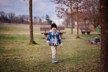 Kışın giyinmiş küçük bir çocuk çimenli bir parkta yürüyor, mavi bir sırt çantası takıyor ve açık alanı keşfediyor..