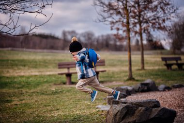 Kış kıyafetleri içinde enerjik bir çocuk parktaki kayaların arasına atlıyor. Üzerinde mavi bir roket çantası var..