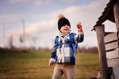 Kışlık giysiler içindeki neşeli çocuk dışarıda tahta bir oyun alanının yanında küçük taşlar tutarken gülümsüyor..