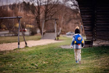 Kışın giysi giymiş küçük bir çocuk çimenli bir parkta neşeli bir şekilde koşar. Üzerinde oyuncak ayıyla birlikte bir roket çantası, güneş batarken..