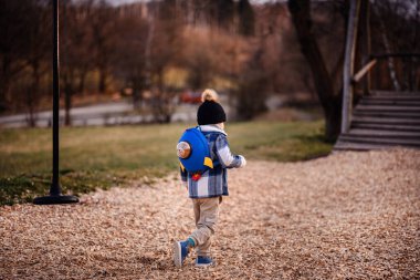 Kışlık giysiler içinde küçük bir çocuk ve mavi bir roket sırt çantası ahşap kırıntıları üzerinde yürür, geride ip salıncak bırakır.