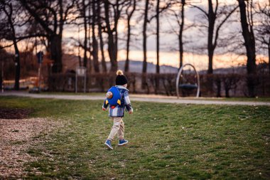Kışın giysi giymiş küçük bir çocuk çimenli bir parkta neşeli bir şekilde koşar. Üzerinde oyuncak ayıyla birlikte bir roket çantası, güneş batarken..