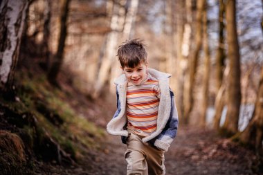 Ekose ceketli gülümseyen bir çocuk güneşli bir günde manzaralı bir orman yolunda koşar..
