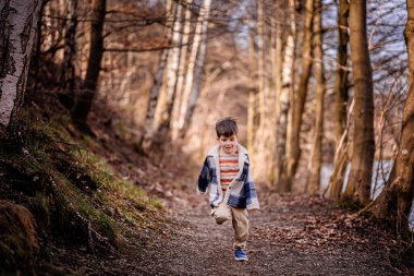 Ekose ceketli gülümseyen bir çocuk güneşli bir günde manzaralı bir orman yolunda koşar..