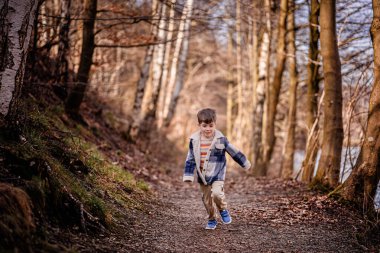 Ekose ceketli gülümseyen bir çocuk güneşli bir günde manzaralı bir orman yolunda koşar..