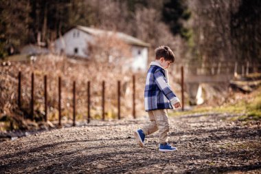 Mavi ekose ceketli genç bir çocuk kırsal ve ağaçlık bir alanda çakıllı bir yolda düşünceli bir şekilde yürüyor..