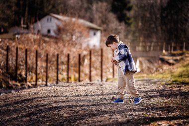 Mavi ekose ceketli neşeli genç bir çocuk taşralı bir kırsal kesimde çakıl yolunda taşlarla oynuyor..