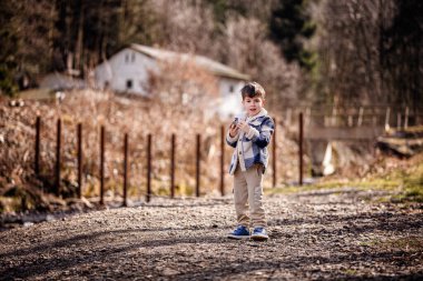 Mavi ekose ceketli neşeli genç bir çocuk taşralı bir kırsal kesimde çakıl yolunda taşlarla oynuyor..