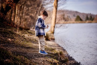 Mavi ekose ceketli bir çocuk güneşli bir öğleden sonra sakin bir göl kıyısında taş sektirmekten hoşlanır..