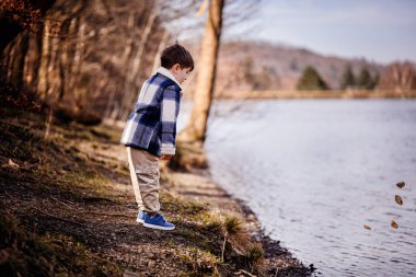 Mavi ekose ceketli bir çocuk güneşli bir öğleden sonra sakin bir göl kıyısında taş sektirmekten hoşlanır..