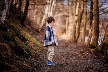 Mavi ekose ceketli bir çocuk ağaçlarla kaplı huzurlu bir orman yolunda yürüyor..