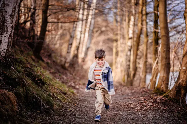 Ekose ceketli gülümseyen bir çocuk güneşli bir günde manzaralı bir orman yolunda koşar..