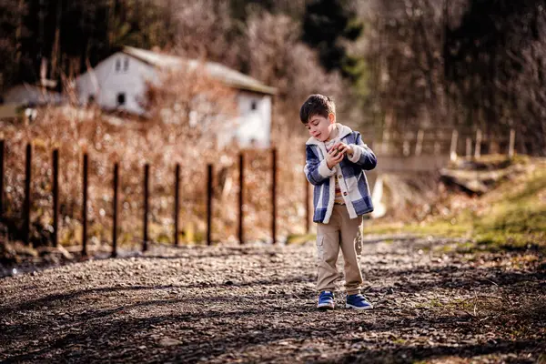 Mavi ekose ceketli neşeli genç bir çocuk taşralı bir kırsal kesimde çakıl yolunda taşlarla oynuyor..