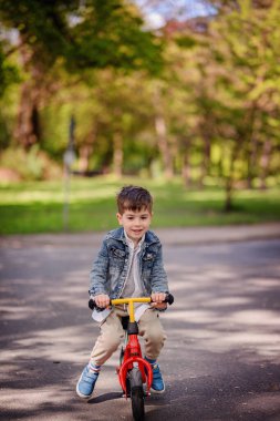 Güneşli bir günde, yeşil bir parkta, gölgeli bir yolda kırmızı dengeli bir bisiklete binen ve açık havanın tadını çıkaran genç bir çocuk..