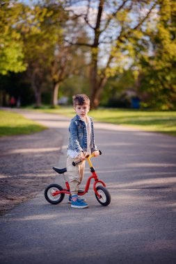 Güneşli bir günde, yeşil bir parkta, gölgeli bir yolda kırmızı dengeli bir bisiklete binen ve açık havanın tadını çıkaran genç bir çocuk..