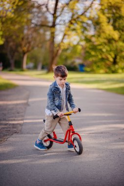 Güneşli bir günde, yeşil bir parkta, gölgeli bir yolda kırmızı dengeli bir bisiklete binen ve açık havanın tadını çıkaran genç bir çocuk..