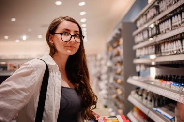 Gözlüklü genç bir kadın iyi aydınlatılmış bir sanat mağazasında raflara ve malzemelere dikkatlice bakıyor..