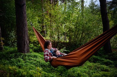 Bir anne, uyuyan oğlunu yemyeşil ormanlarla çevrili turuncu bir hamakta tutar. Birlikte sakin ve hassas bir anın tadını çıkarır..
