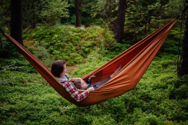 Bir anne ve oğlu, yemyeşil orman ve uzun ağaçlarla çevrili turuncu bir hamakta huzurlu bir anı paylaşıyorlar..