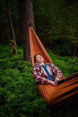 Ekose ceketli bir kadın, yemyeşil ormanlarla çevrili turuncu bir hamakta gözleri kapalı dinleniyor..