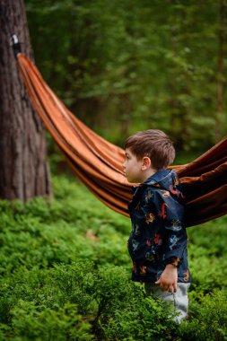 Renkli ceketli genç bir çocuk turuncu bir hamağın yanında duruyor, merak ve merakla ormana bakıyor..