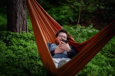 Gözlüklü ve sakallı bir adam ormanın içinde turuncu bir hamakta uzanıyor, açık havada huzurlu bir anda akıllı telefonuna odaklanıyor..