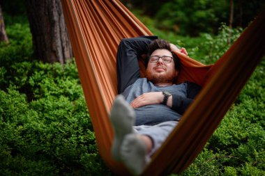 Gözlüklü rahat bir adam turuncu bir hamakta uzanıyor, yemyeşil ve doğada sakin bir anın tadını çıkarıyor..