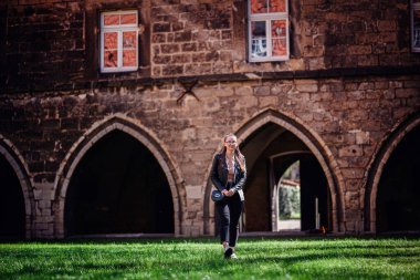 Gözlüklü ve deri ceketli şık genç bir kadın kemerli kapılı orta çağ taş yapısının önünde çimlerin üzerinde yürüyor..