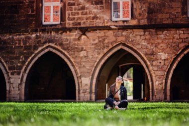 Gözlüklü şık bir kadın ve deri ceketli yeşil çimlerin üzerinde oturuyor. Ortaçağ taş yapısının önünde kemerli girişleri olan..