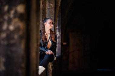 Gözlüklü ve deri ceketli kadın sakin bir şekilde tarihi bir koridorda oturuyor, doğal ışıkla hafifçe aydınlatılıyor..