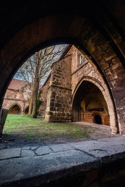 Koyu taştan bir kemerin içinden bakınca kemerli ortaçağ mimarisi ve sarmaşık kaplı duvarlarla çevrili çimenli bir avlu görünüyor..