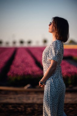 Şık bir kadın gün batımında çiçek açan bir lale tarlasında dikiliyor, polka noktalı bir elbise ve açık renk bir gömlek giyiyor, sıcak altın ışığa bakıyor..