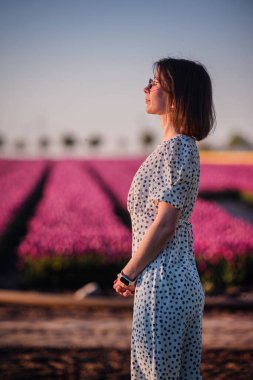 Şık bir kadın gün batımında çiçek açan bir lale tarlasında dikiliyor, polka noktalı bir elbise ve açık renk bir gömlek giyiyor, sıcak altın ışığa bakıyor..