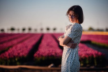 Güneş gözlüklü ve benekli elbiseli zarif kadın gün batımında canlı pembe lale tarlasında güven içinde poz veriyor..