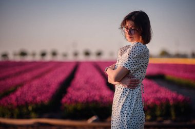 Güneş gözlüklü ve benekli elbiseli zarif kadın gün batımında canlı pembe lale tarlasında güven içinde poz veriyor..