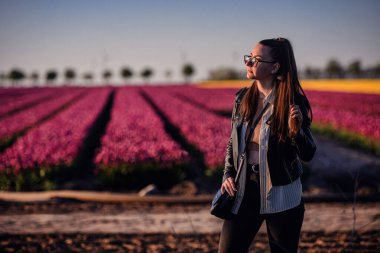 Deri ceketli, gözlüklü şık bir kadın altın akşam güneşi altında canlı lale sıralarının önünde poz veriyor..