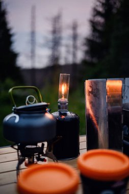 Yanan bir gaz lambası, ormanlık bir alanda alacakaranlıkta, bir çaydanlık ve ahşap bir masada yemek takımının yanında parlar..