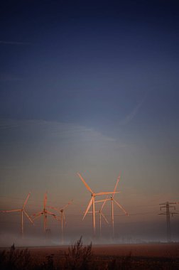 Tall wind turbines stand in a misty field at dawn, generating clean energy.