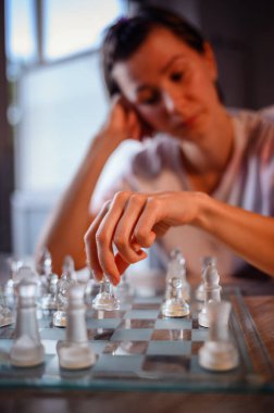 Hareket sırasında buzlu cam bir satranç taşını kaldıran bir elin yakın çekimi, arka planda bulanık dalgın bir oyuncu.